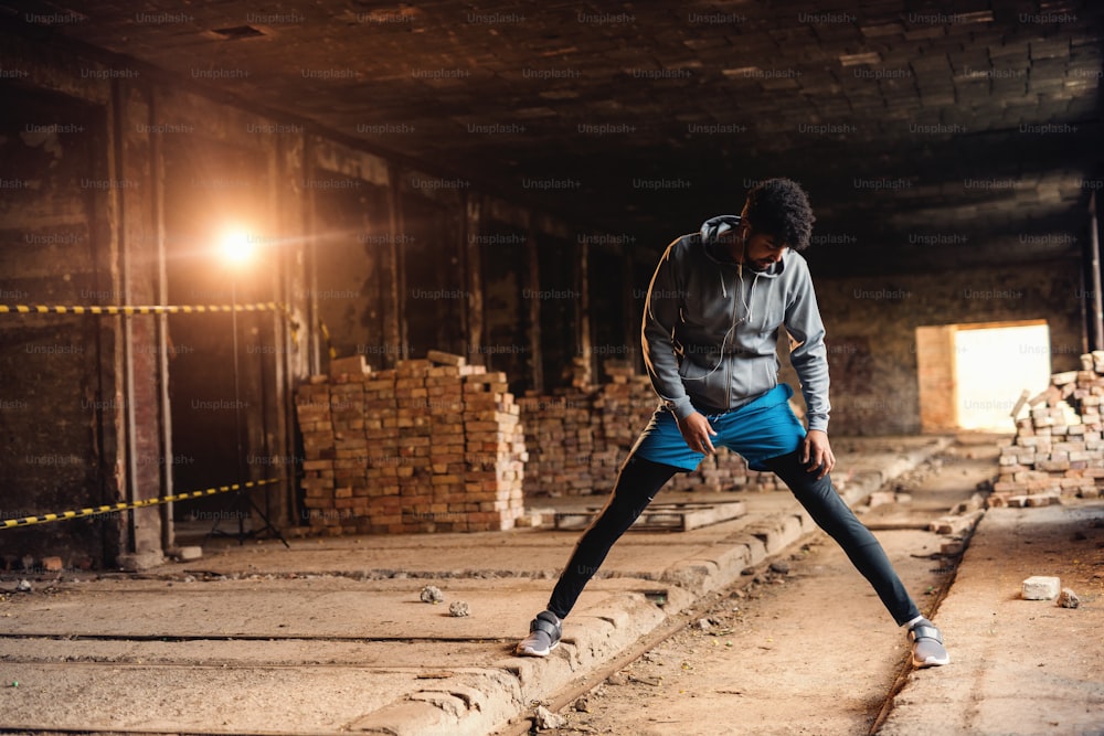 Hombre deportivo afroamericano en ropa deportiva estirando las piernas en una vieja fábrica de ladrillos. Concepto de estilo de vida saludable.