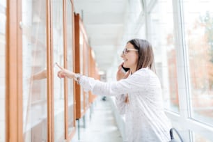 Smiling collage girl with brown hair and eyeglasses using smart phone for calling her colleague to report who passed exams, pointing with finger at noticeboard. University building interior.