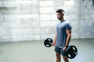 Caucasian serious bodybuilder standing and holding barbell. In background wall.