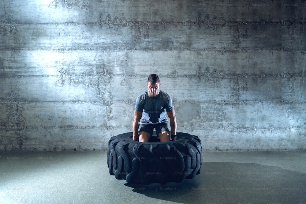 Homem musculoso forte em pneu de levantamento de roupa esportiva enquanto está em pé na frente da parede no ginásio ginásio.