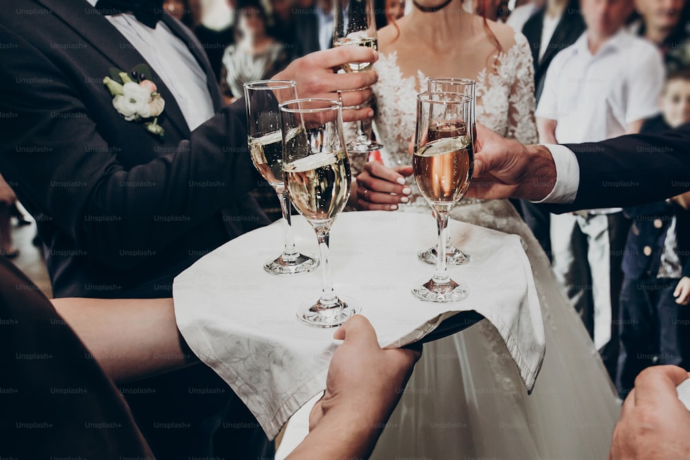 쟁반에 샴페인 잔, 안경을 들고 건배하는 손, 결혼식 축하. 가족 응원과 함께 세련 된 행복한 신혼 부부. 텍스트 공간. 호화로운 결혼식 피로연