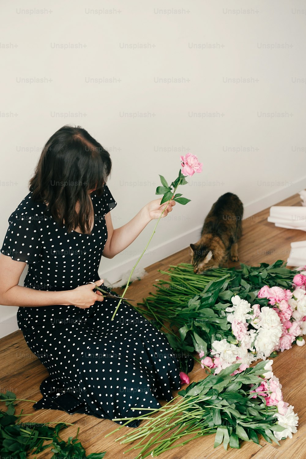 Beautiful girl in vintage dress holding scissors and cutting stems of pink and white peonies. Happy stylish woman arranging peony flowers with cat in room. Florist in studio.