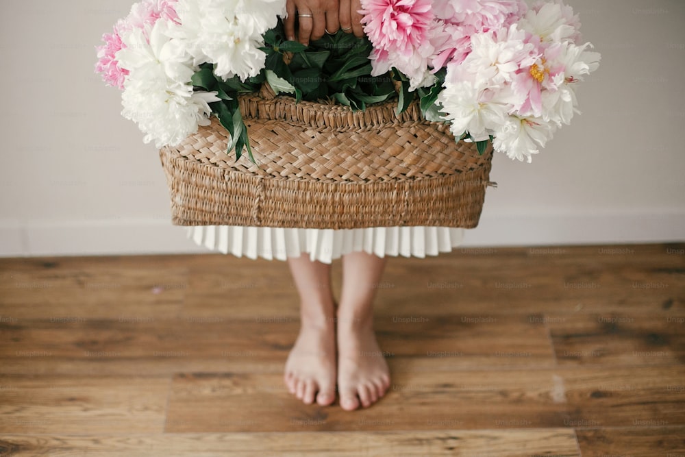 Boho girl holding pink and white peonies in rustic basket and standing barefoot on wooden floor. Stylish hipster woman in bohemian floral dress with peony flowers. International Womens Day