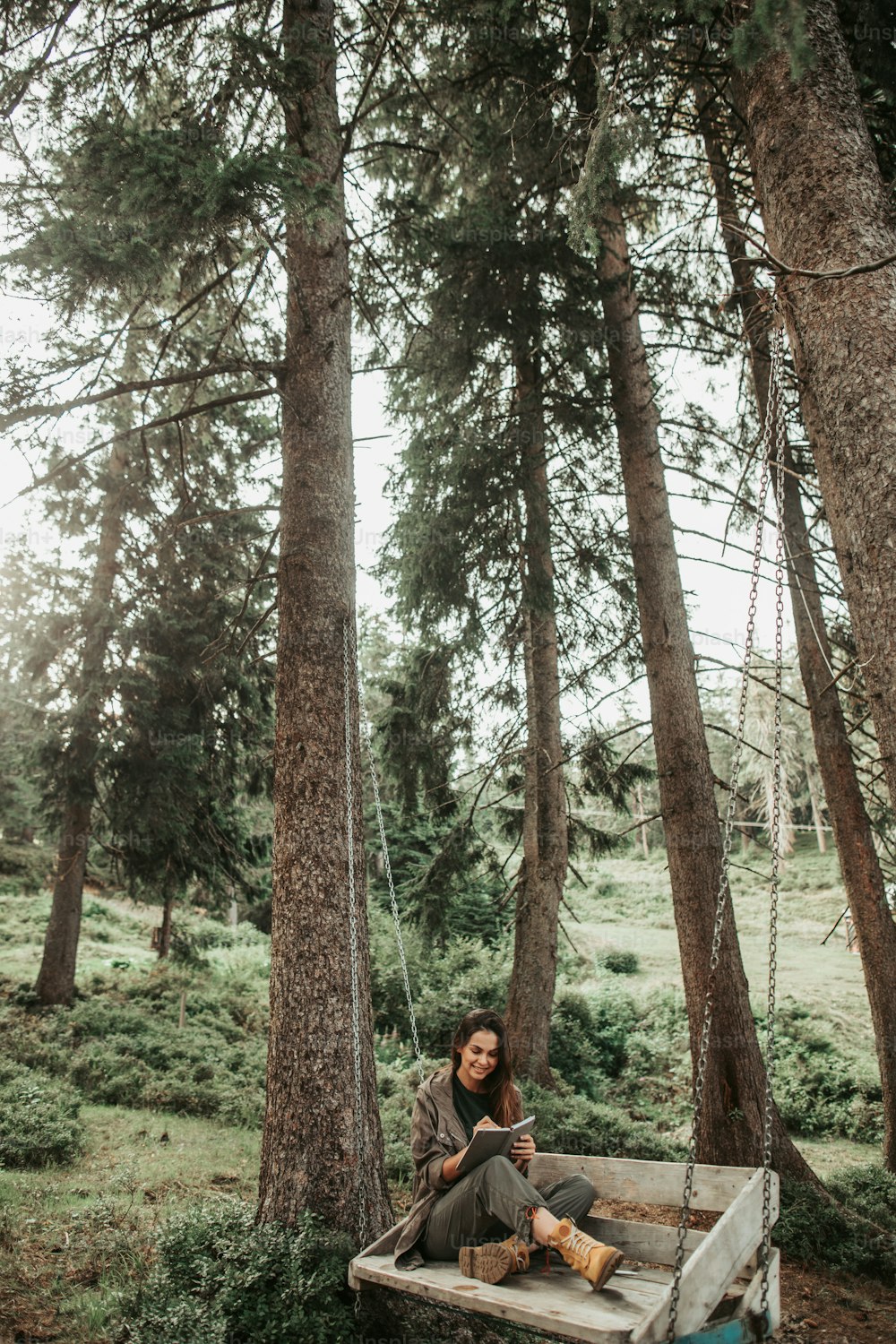 숲에서 영감을 얻는다. 노트북에 쓰고 웃고 있는 매력적인 젊은 여성의 초상화. 그녀는 키 큰 나무들로 둘러싸인 숲에서 쉬고 있다