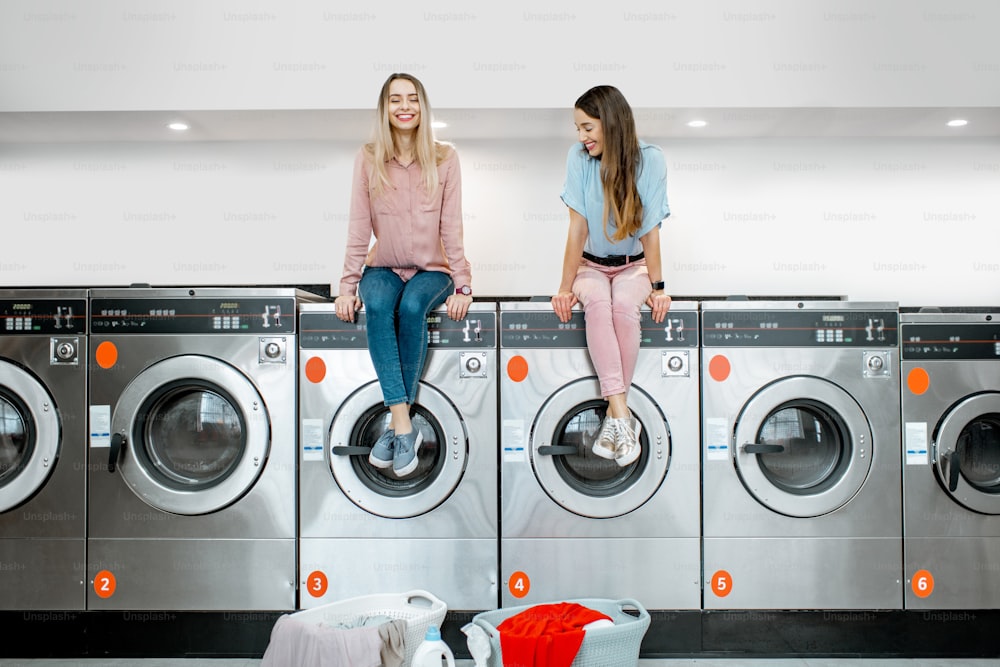 Two cheerful girlfriends waiting for washing while sitting on the washing machines in the self-service laundry