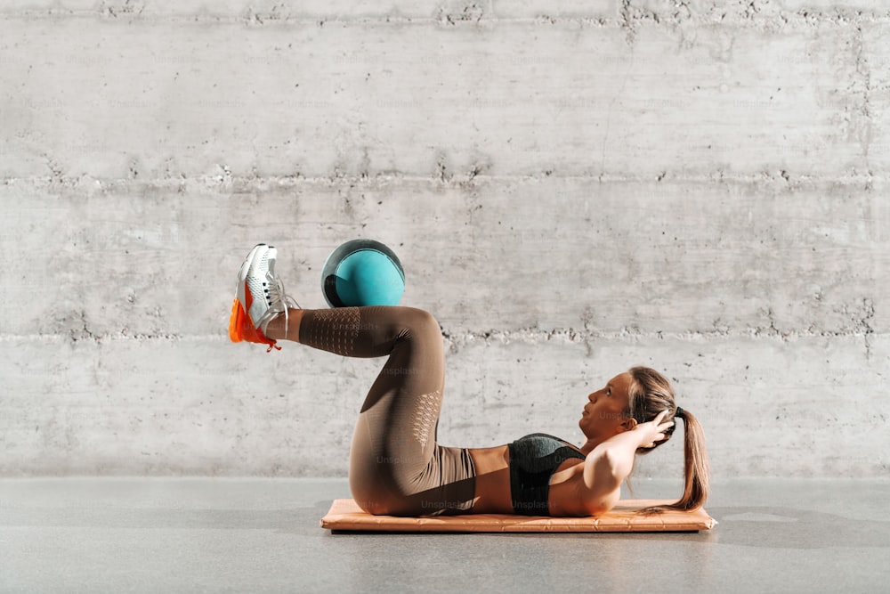 ポニーテールで、灰色の壁の前のマットの上でボールで腹筋をしているスポーツウェアを着た筋肉質の強力な女性の側面図。
