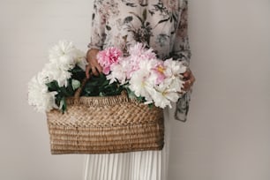 Boho girl holding pink and white peonies in rustic basket. Stylish hipster woman in bohemian floral dress gathering peony flowers. International Womens Day.  Wedding decor and arrangement