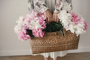 Boho girl holding pink and white peonies in rustic basket. Stylish hipster woman in bohemian floral dress gathering peony flowers. Happy mothers day. International Womens Day.