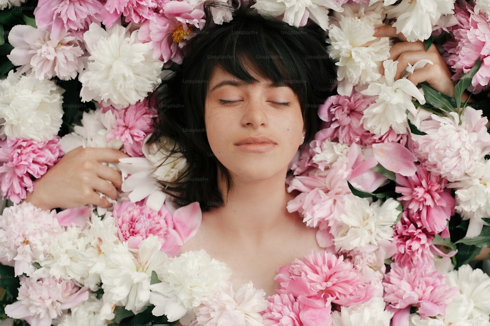 Beautiful brunette girl in many pink and white peonies. Happy boho woman portrait with peony flowers, top view. Creative floral photo. Aroma scent concept. International Womens Day