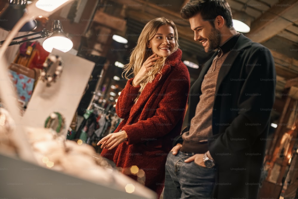 Good quality handmade imitation jewelry. Happy young joyful couple choosing handmade imitation jewelry at small street market. Autumn season, blond haired woman looking at her boyfriend at street market