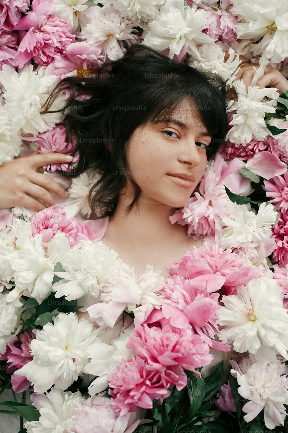 Beautiful brunette girl lying in many pink and white peonies. Boho woman portrait with natural makeup in peony flowers. Creative floral photo. Aroma scent concept. International Womens Day