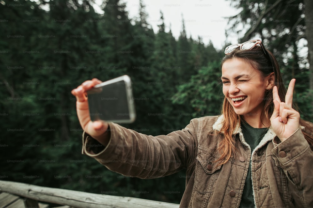 Divertirsi nel bosco. Ritratto di giovane donna affascinante che mostra il gesto di pace mentre scatta foto con cellulare. Fa l'occhiolino e sorride