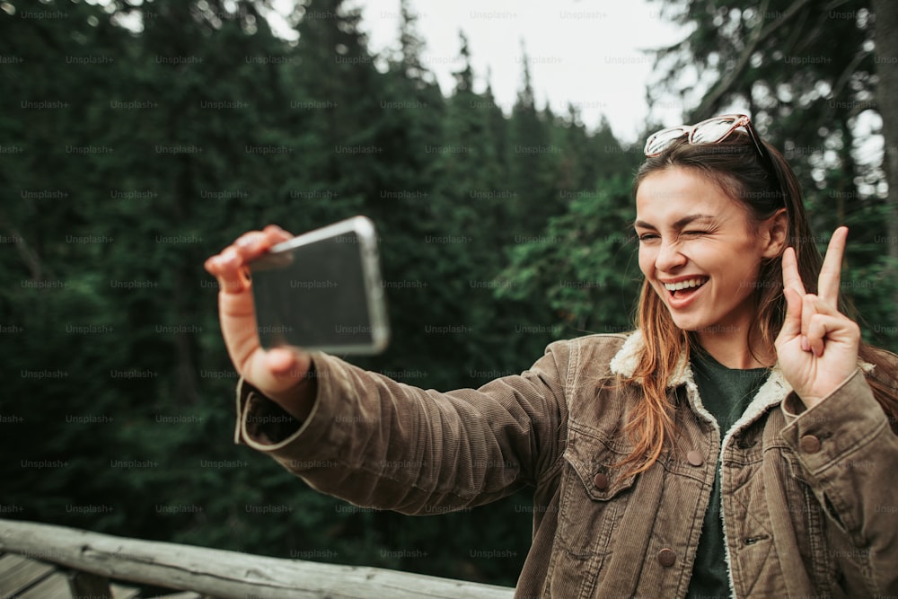 Divirtiéndose en el bosque. Retrato de una joven encantadora que muestra un gesto de paz mientras toma una foto con el teléfono celular. Ella guiña un ojo y sonríe