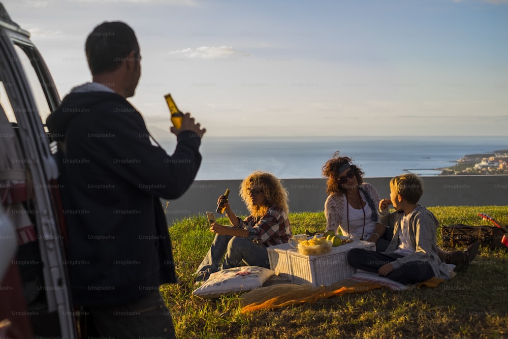 picnic and travel concept for family caucasian people enjoying the sunset sitting in the meadow and drinking beer together - children eating having fun with mother - ocean view and vacation lifestyle time