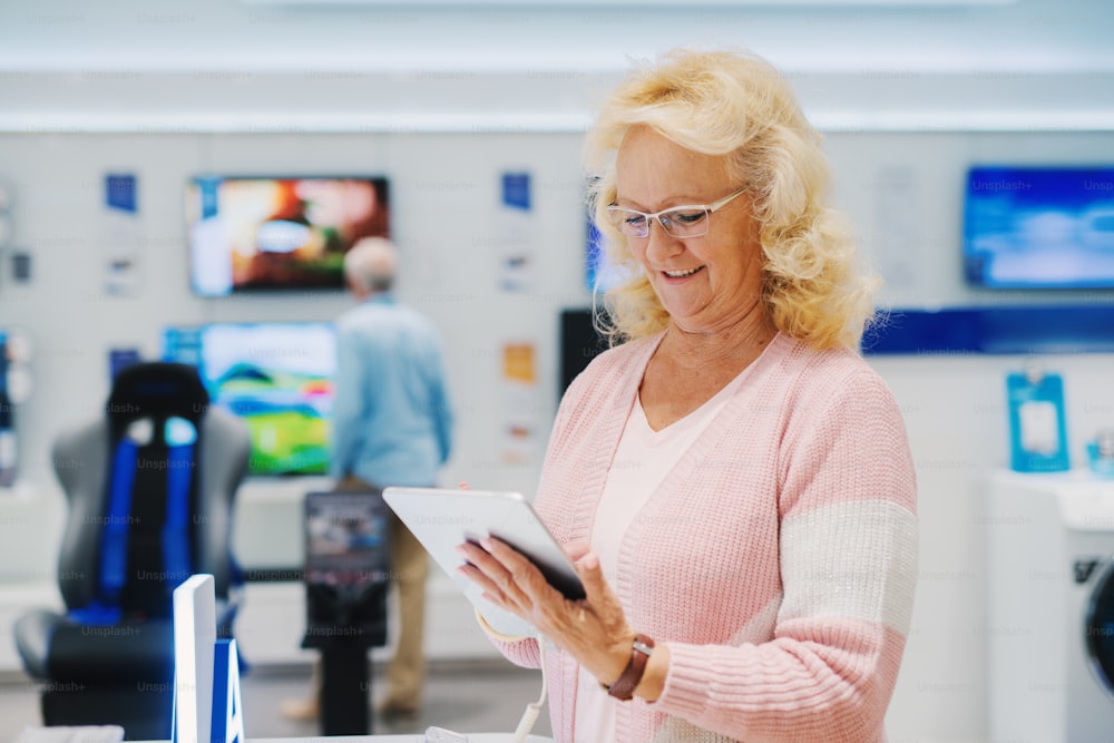 Mujer mayor caucásica sonriente con cabello rubio y anteojos probando una tableta mientras está de pie en una tienda de tecnología.