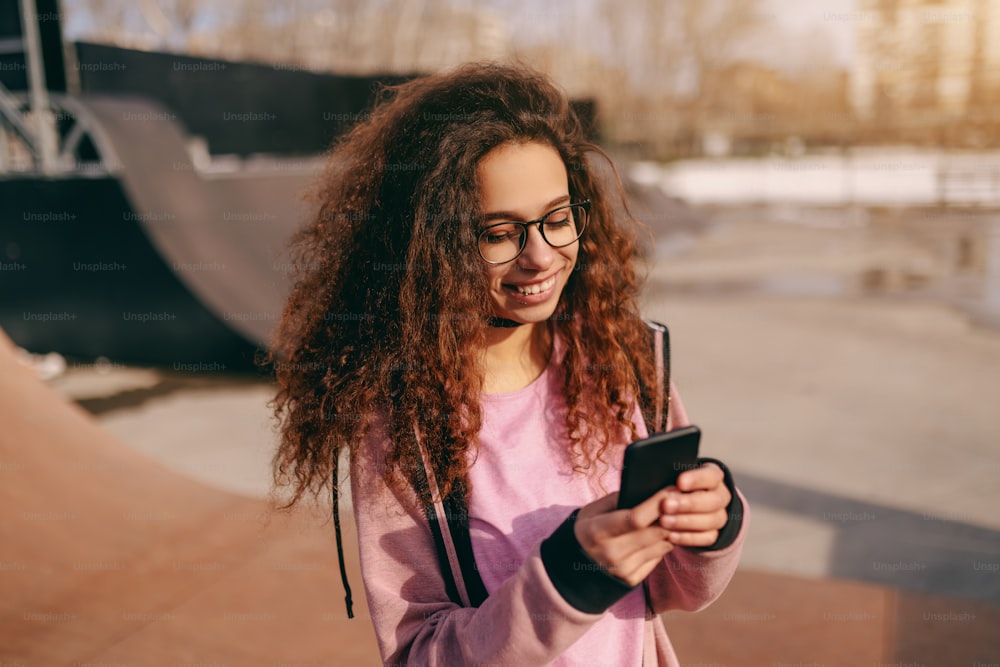 Nahaufnahme eines hübschen gemischtrassigen Teenager-Hipster-Mädchens mit zahnigem Lächeln mit Smartphone, während es im Skatepark steht.