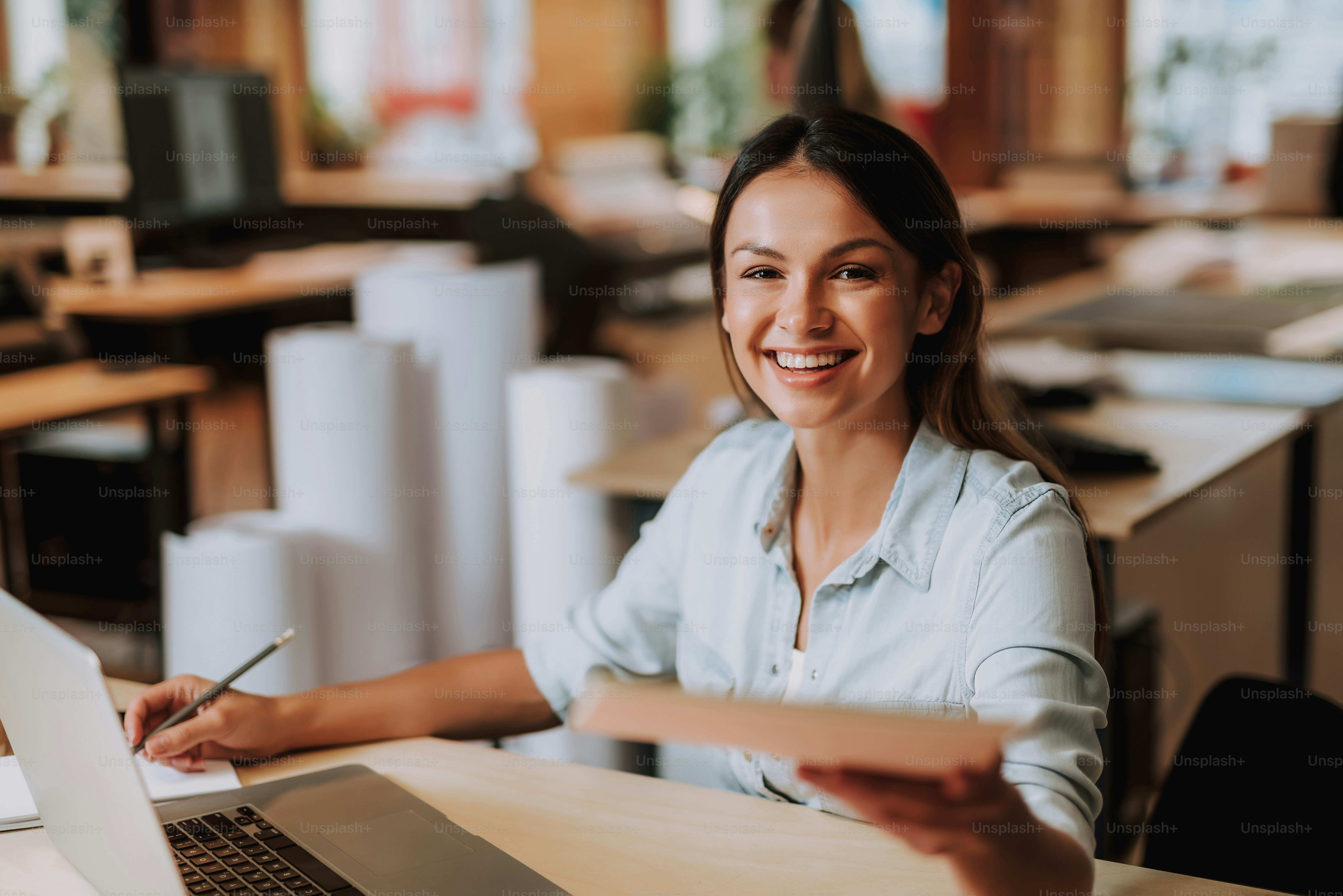 Portrait of beautiful woman making notes and holding papers while sitting at office desk. She is looking at camera and smiling