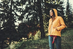 I am ready for adventures. Portrait of charming girl in yellow jacket enjoying sunny day in coniferous wood. She is looking away with smile and keeping hands in pockets