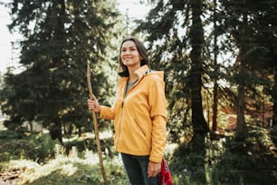 Brave hiker. Portrait of cute girl in yellow jacket with tree branch in her hand spending time in sunny coniferous wood. She is looking away with smile