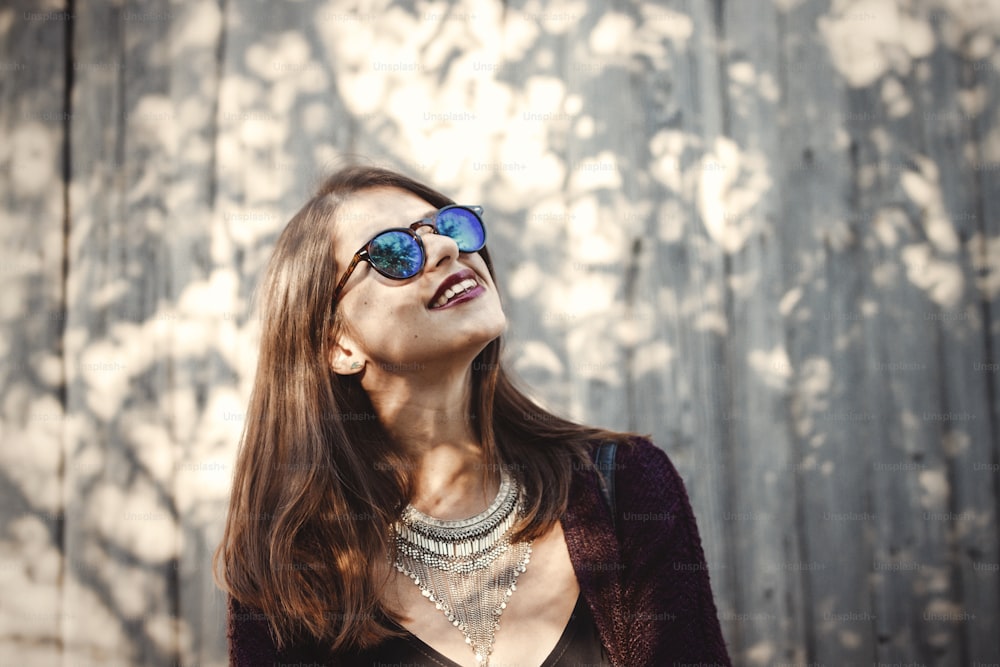 Stylish hipster girl smiling in sunny street on background of wooden wall. Boho girl in cool outfit and sunglasses posing in sunlight and shadow. Space for text. Summer vacation and travel