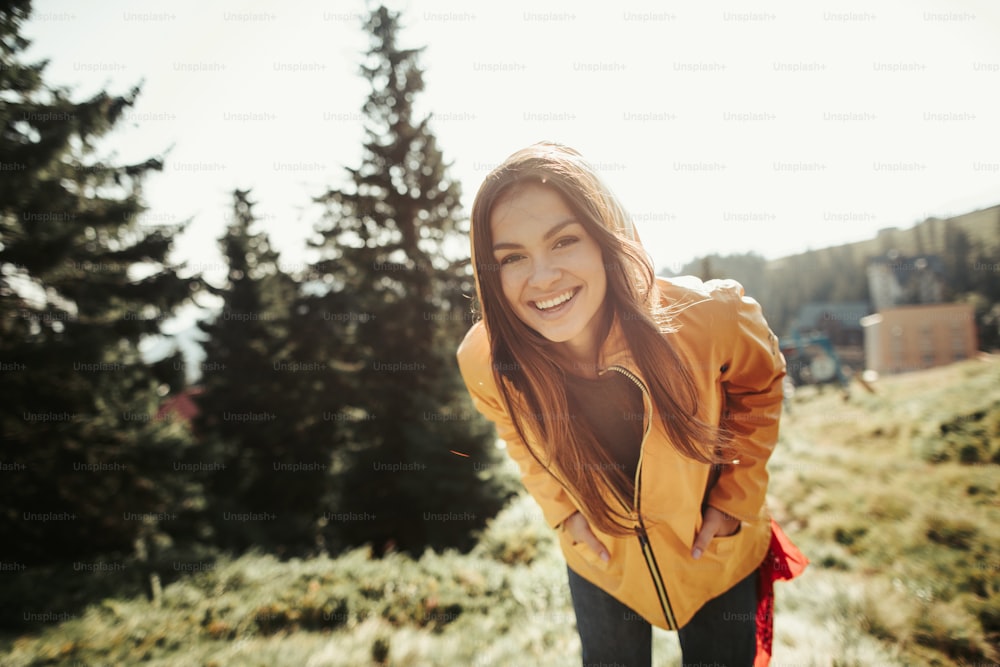 I have a wonderful day. Portrait of cheerful young lady in yellow jacket standing in coniferous wood and keeping hands in pockets. She is bending down a little and looking at camera with happy smile