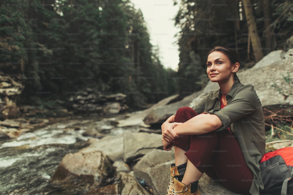 Mulher jovem amigável sentada nas rochas do rio da montanha enquanto desfruta de belas vistas