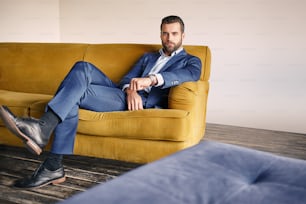 Good looking young businessman in a stylish suit is relaxing on sofa and looking at camera. Business look.