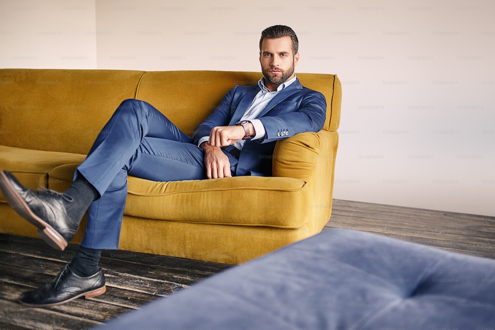 Un giovane uomo d'affari di bell'aspetto in un abito elegante si rilassa sul divano e guarda la macchina fotografica. Aspetto aziendale.