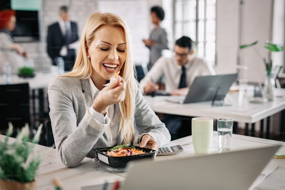 Lächelnde Geschäftsfrau, die bei der Arbeit gesund isst und Gemüsesalat zum Mittagessen hat.