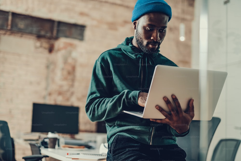 Ruhiger, attraktiver junger Mann in Freizeitkleidung, der allein im Büro sitzt und einen modernen Laptop benutzt, während er ihn in den Händen hält