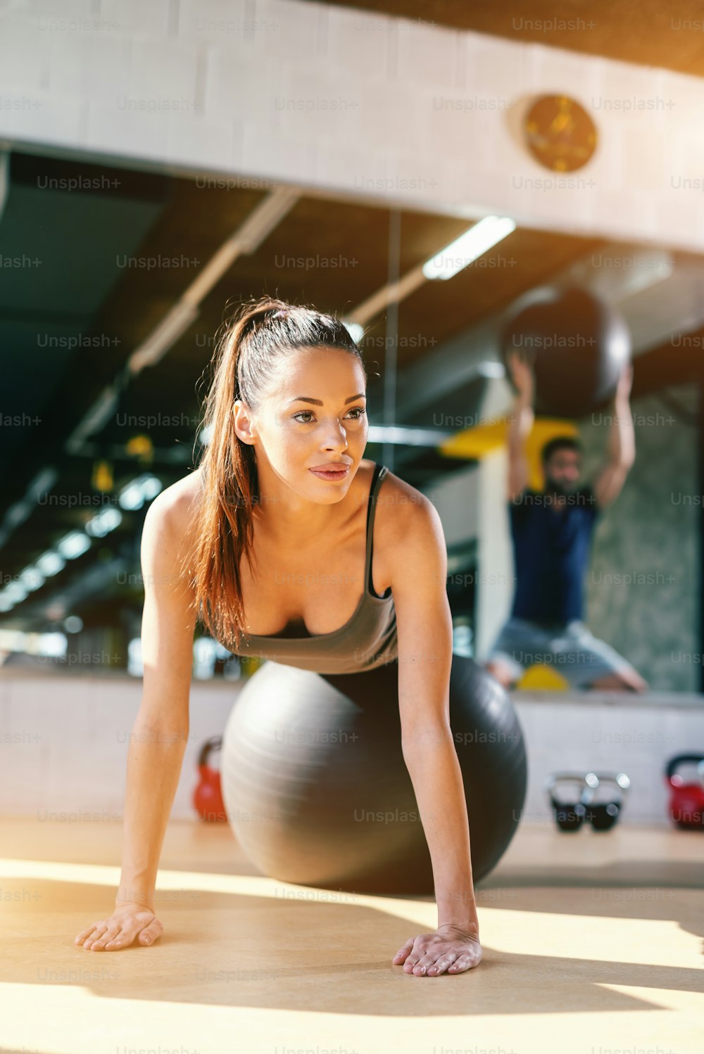 Close up of beautiful Caucasian woman with ponytail and long brown hair doing exercises with pilates ball in gym. In background reflection of man with pilates ball.