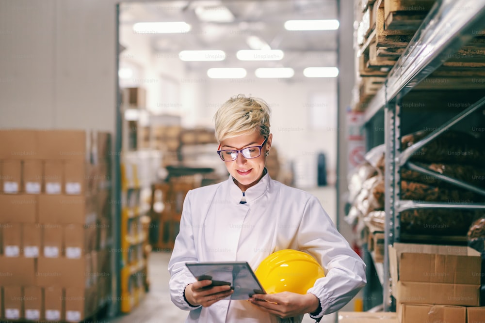 Young Caucasian blonde female employee with protective helmet under armpit using tablet while standing in warehouse.