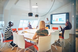 Gerente discutiendo gráficos en un monitor durante una reunión con un grupo diverso de colegas sentados juntos dentro de una oficina de vidrio