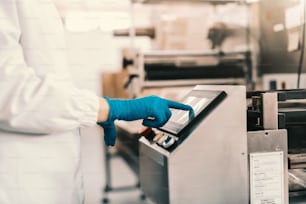 Close up of young female employee in sterile uniform and blue rubber gloves turning on packing machine while standing in food factory.