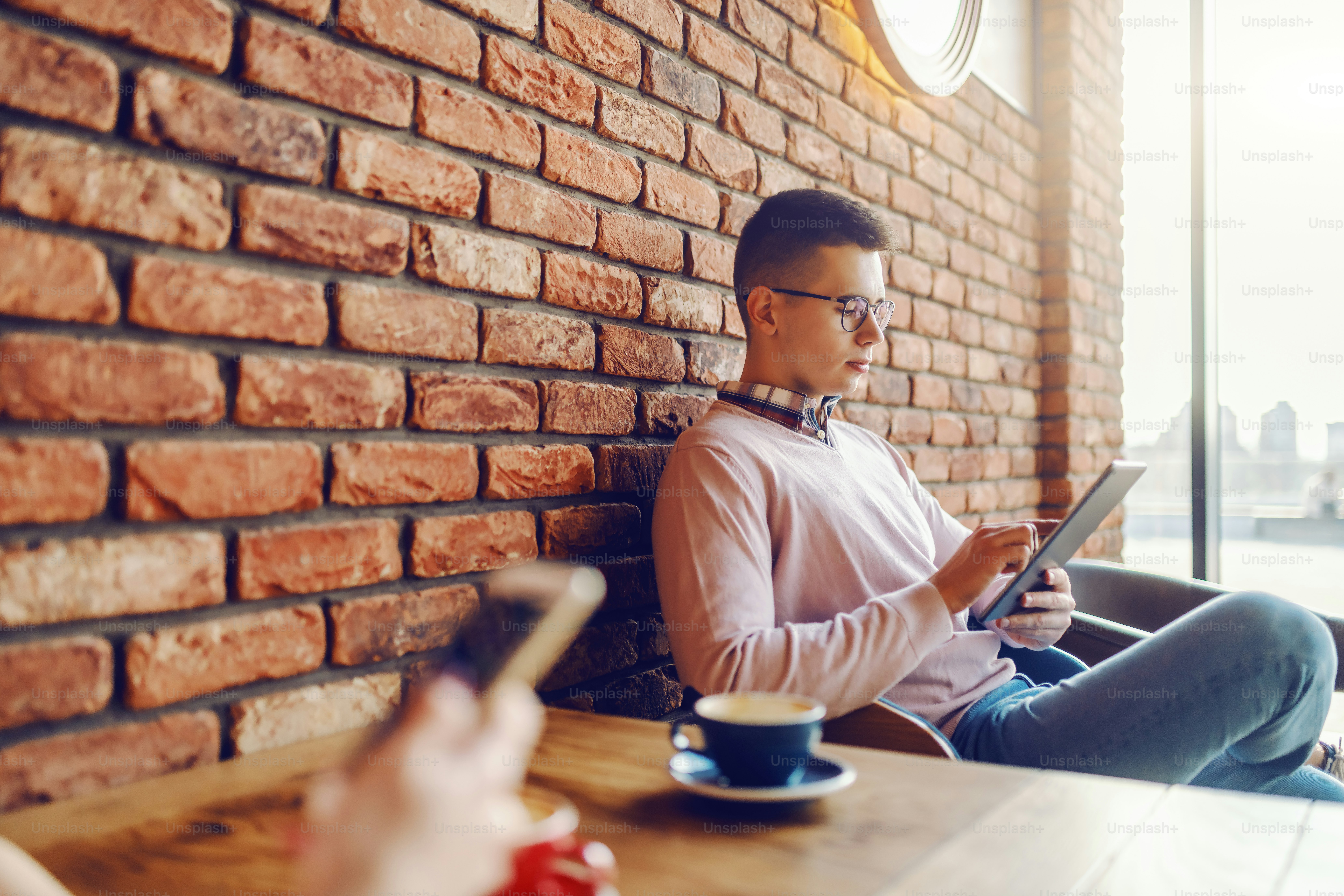 Pria muda Kaukasia dengan ekspresi wajah yang serius dan berpakaian kasual yang cerdas menggunakan tablet. Di latar depan wanita menggunakan ponsel pintar. Fokus selektif pada manusia.