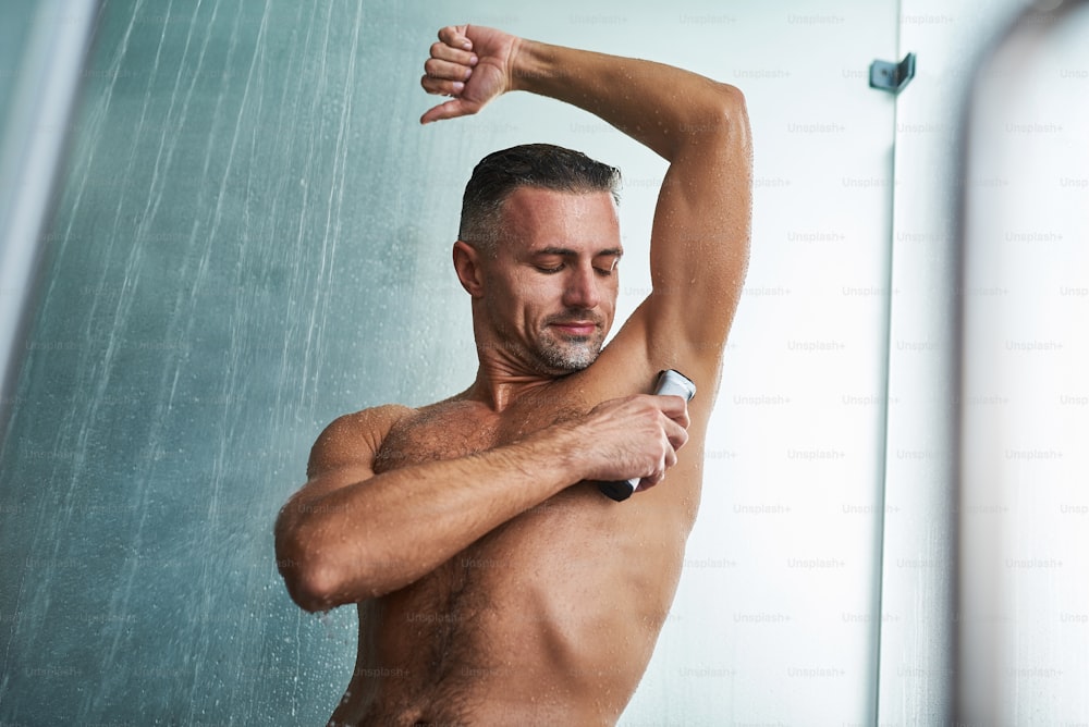 Waist up portrait of handsome gentleman with wet body using electric razor to remove hair from underarm