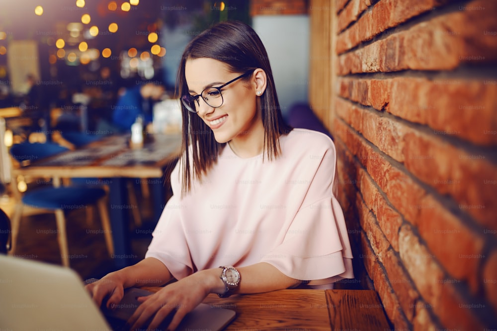 Attractive brunette with eyeglasses using laptop while sitting inn cafeteria. In background bokeh effect.