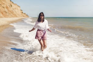 Happy young woman in white shirt and sunglasses running and having fun with waves on sunny beach. Hipster slim girl relaxing and smiling at sea. Summer vacation concept. Copy Space. Joyful moment