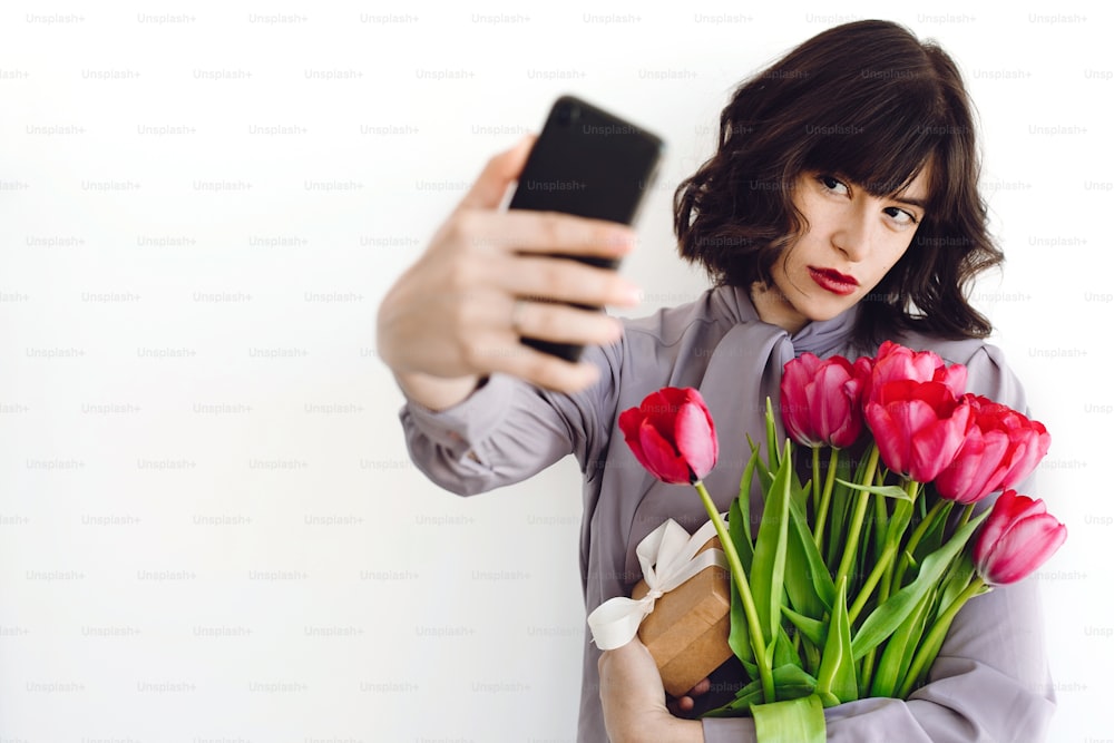 Beautiful brunette girl holding bouquet of tulips and gift box and taking selfie on white background indoors, space for text. Stylish young woman with flowers and phone. Happy mothers day