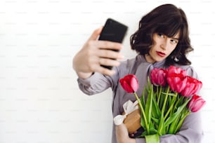 Belle fille brune tenant un bouquet de tulipes et une boîte-cadeau et prenant un selfie sur fond blanc à l’intérieur, espace pour le texte. Jeune femme élégante avec des fleurs et un téléphone. Bonne fête des mères