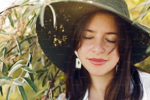 Stylish girl calm portrait in hat posing at olive tree branches in warm sunny light. Happy young boho woman with natural skin relaxing in countryside at sunset. Summer vacation.