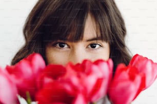 Schönes brünettes Mädchen, das rote Tulpen auf weißem Hintergrund im Haus riecht, Platz für Text. Stilvolles junges Frauenporträt zwischen Tulpen mit attraktiver Optik. Fröhlicher Frauentag