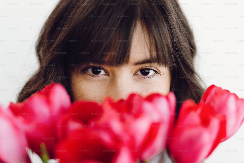 Bella ragazza bruna che sente l'odore dei tulipani rossi su sfondo bianco all'interno, spazio per il testo. Elegante ritratto di giovane donna tra tulipani con aspetto attraente. Buona festa della donna