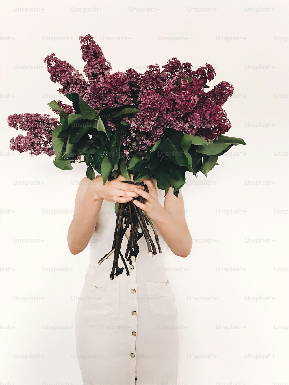 Girl in rustic linen dress holding purple lilac flowers in front of face on white background. Hello spring. Happy Mothers day. Lilac bouquet in woman hands. International womens day