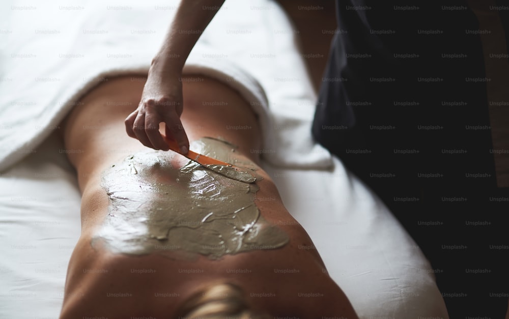 Concept of relaxation and body care. Close up portrait of young woman laying on massage couch while therapist applying healthy spa mud on her back in beauty salon