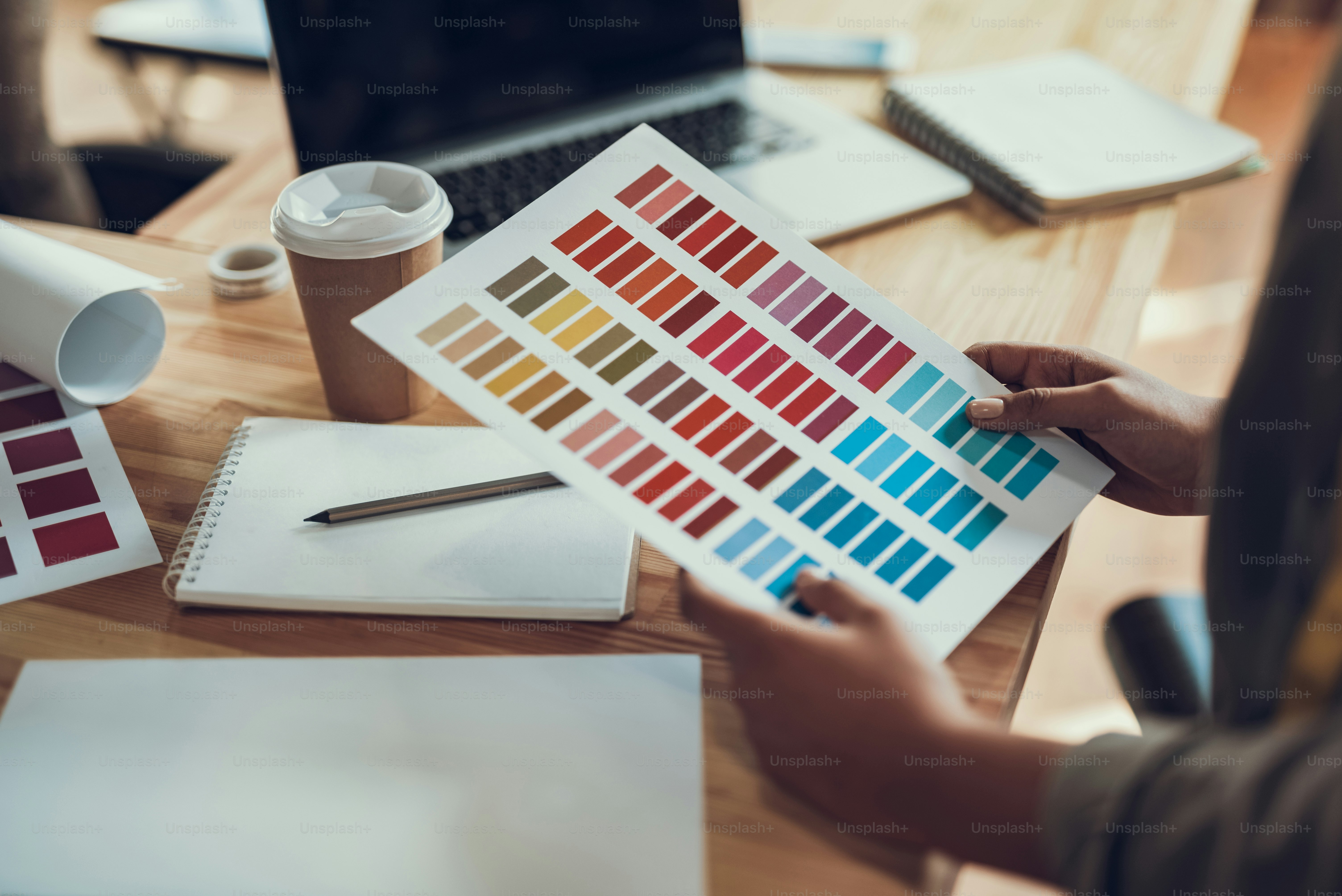 Close up of female hands holding color palette. Girl standing at wooden table with laptop, cup of coffee and stationery