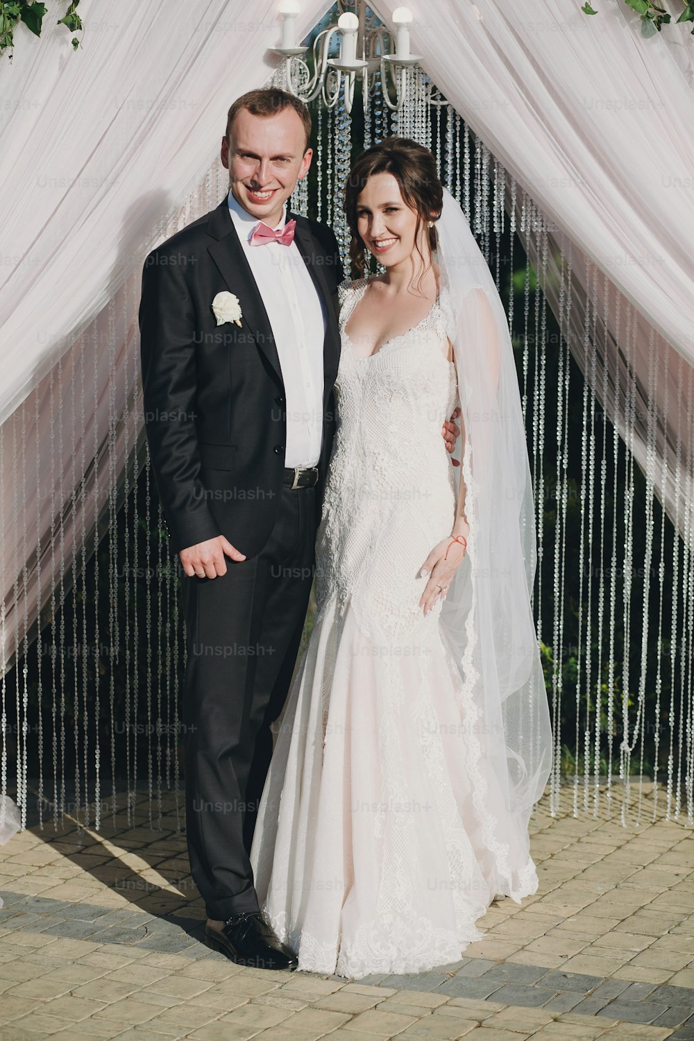 Gorgeous bride and stylish groom posing at stylish wedding arch at wedding reception in restaurant. Luxury wedding couple smiling