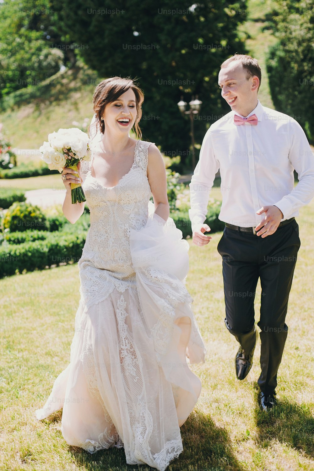 素晴らしいガウンを着たゴージャスな花嫁と、日当たりの良い公園で走って笑うスタイリッシュな花婿。美しい幸せな結婚式のカップルは、時間を楽しんで、屋外で楽しんでいます。ロマンチックなひととき