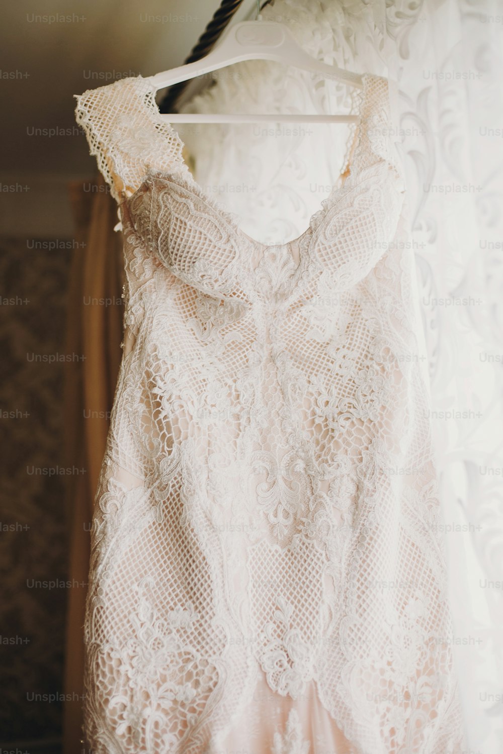 Luxury modern wedding dress hanging at window. Amazing stylish wedding gown with lace floral details, pastel pink color. Wedding salon