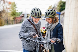 An active senior couple with helmets and electrobikes standing outdoors on a pathway in town, looking at speedometer.